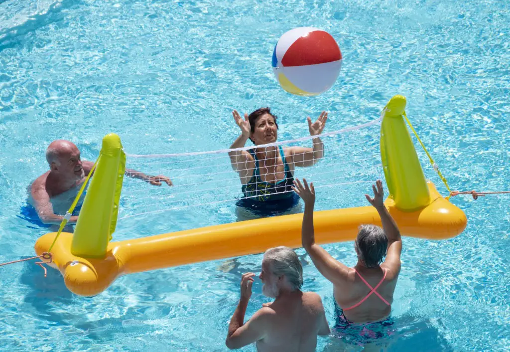 Group of four senior people playing volleyball in the pool with inflatable net and ball. Happiness and smiles shining sun in summer