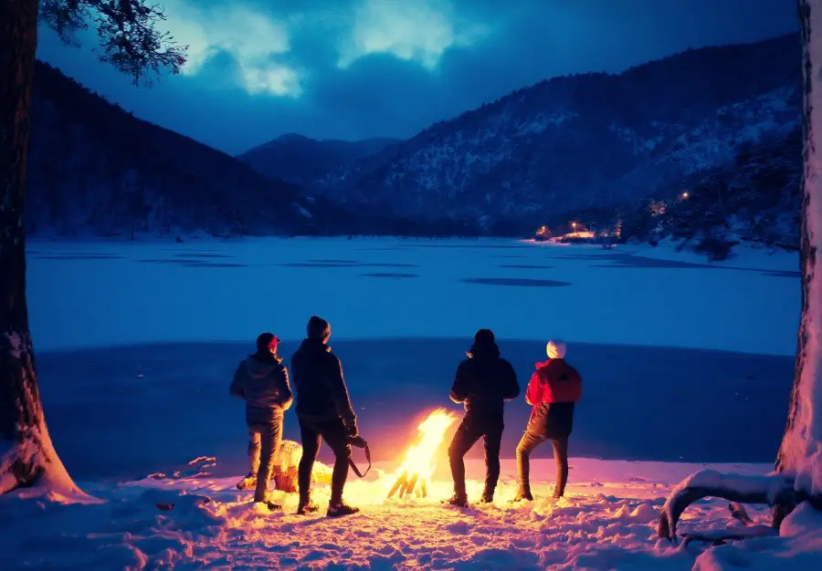 4 people standing around a large campfire beside a mountainside lake in the winter.