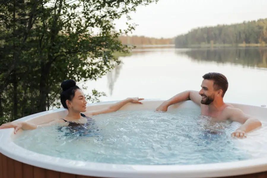 A man and woman relaxing in a hot tub set outdoors beside a lake