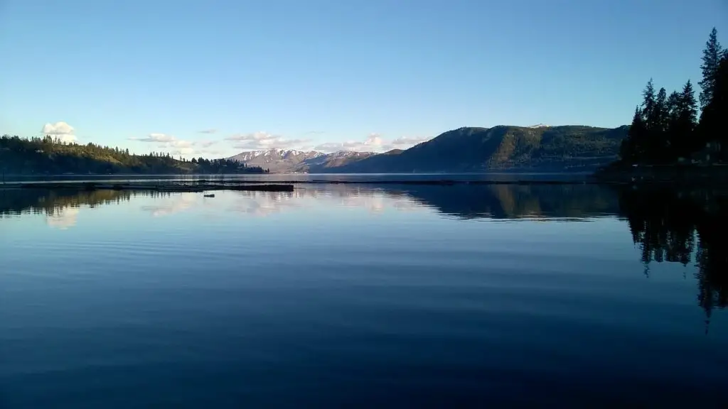 Calm water of Lake Chelan at sunrise with blue sky above and snow-capped mountains far in the distance.