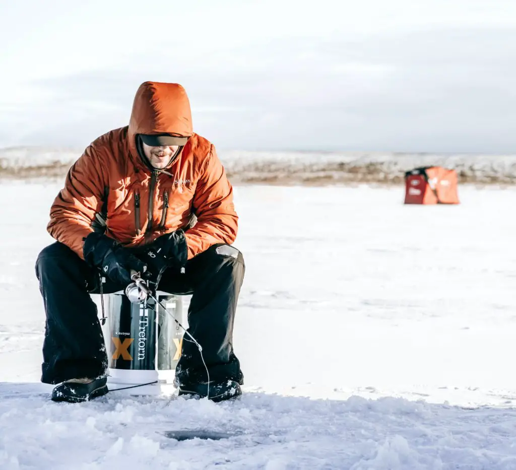 Man bundled up in winter clothes sitting on a bucket on a frozen lake ice fishing. An Ice fishing hut is in the distance