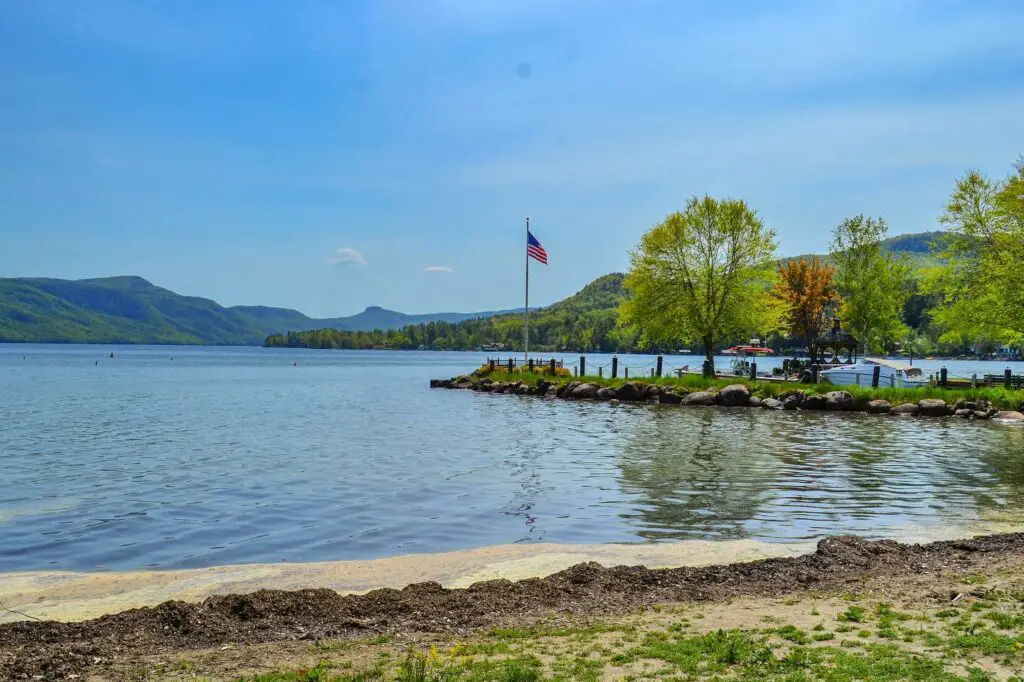 Looking out at Lake George from a small beach. On the right is a small outcropping of land with a tree and American flag flying at full mast. 