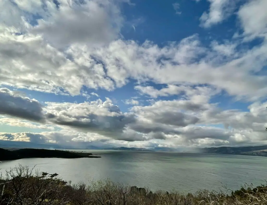 Looking out over clear lake from a grass-covered shoreline. Large white clouds are high in the blue sky.