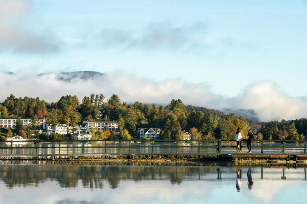 Looking at shoreline from Lake Placid. In the foreground is a dock with three people standing looking at the water. In the background is a treelined hill with large homes along the shoreline. 