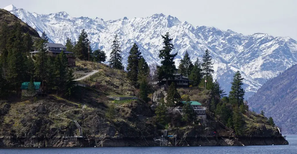 Large rocky cliffs lining Lake Chelan with large homes and pine trees dotting the cliffsides. In the background are large snow-covered mountains. 