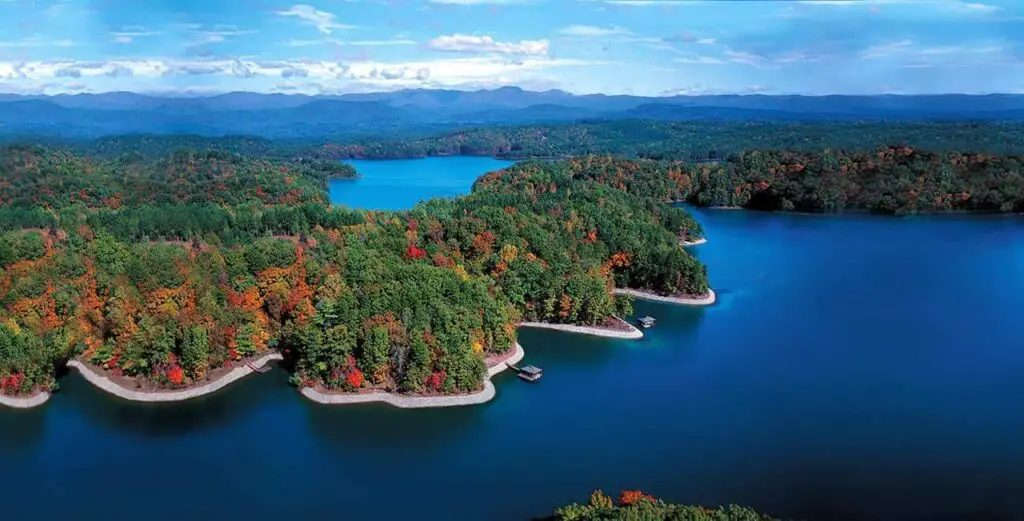 Aerial view of Lake Keowee. A curvy beach-edged shoreline is covered with trees. Docks and boats line the shore. The lake water is deep blue.