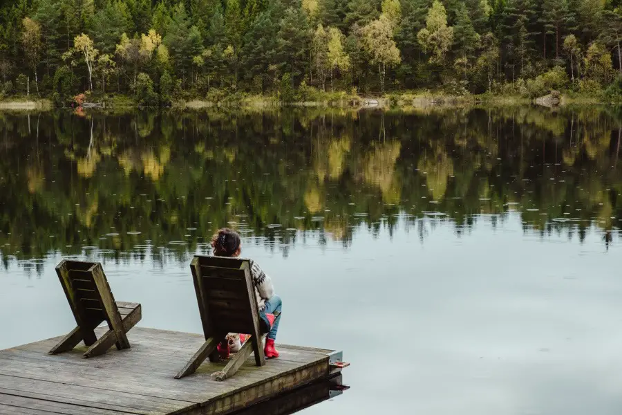 Woman sitting on chair on wooden floating dock looking out on mirror-calm lake surrounded by pine trees.